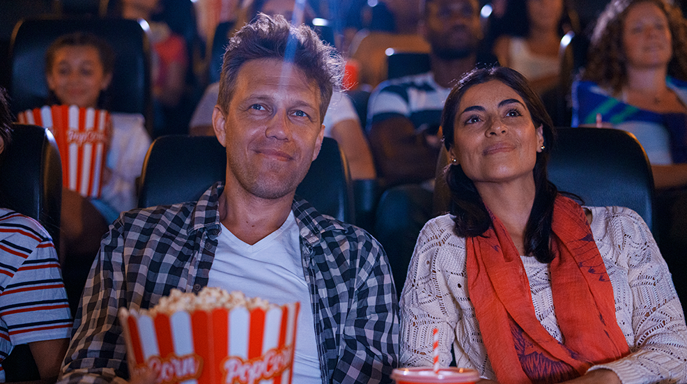 A couple face on sat in a cinema smiling at the screen and holding a large box of popcorn
